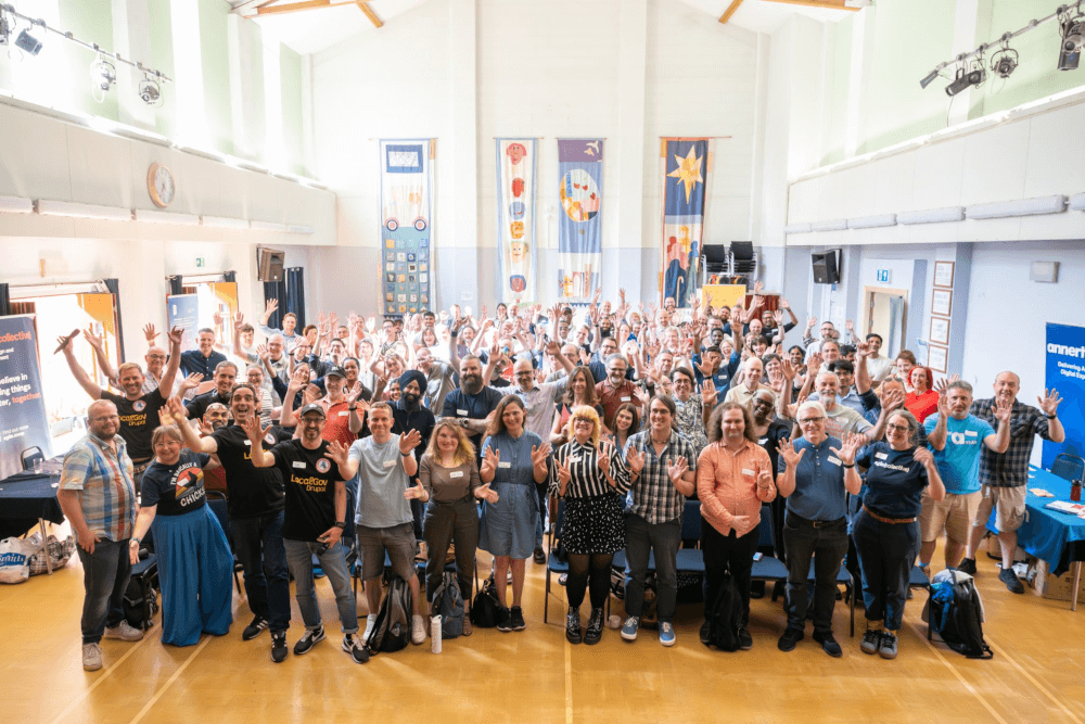 The group photo from LocalGov Drupal Camp 2025. In a room with a high ceiling there are about 100 people in rows who are waving to the camera.